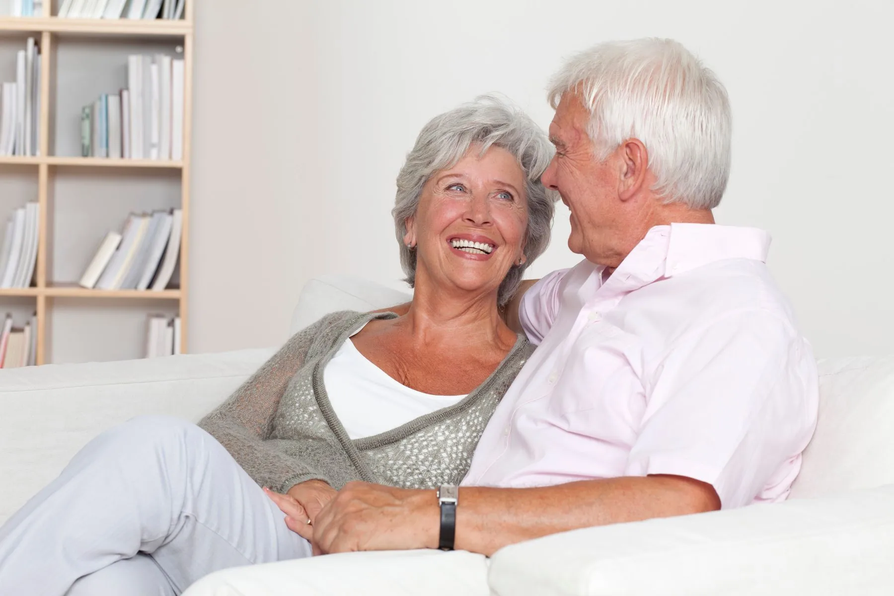 An older couple smiling together on a white sofa in a bright living room